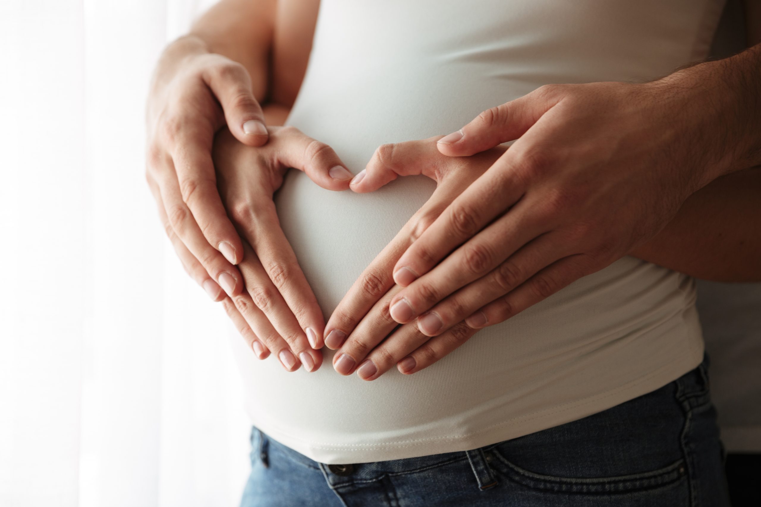 Close up portrait of man's and woman's hands making heart gesture over pregnant belly indoors