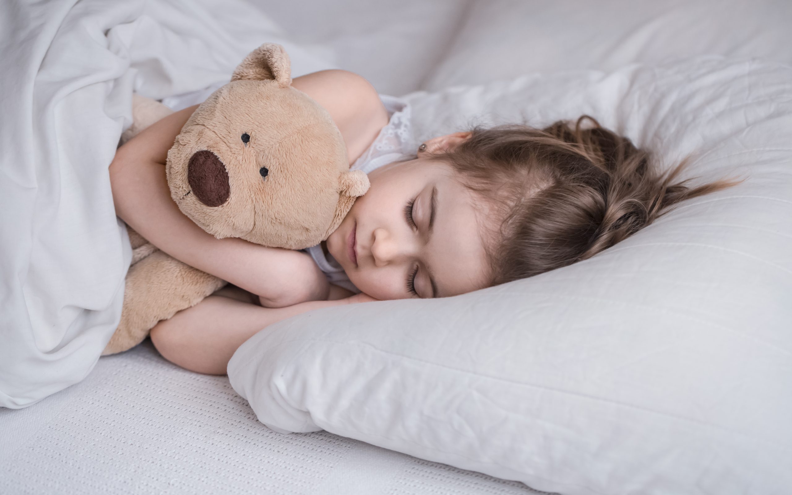 Cute little girl sleeps sweetly in a white cozy bed with a soft bear toy, the concept of children's rest and sleep
